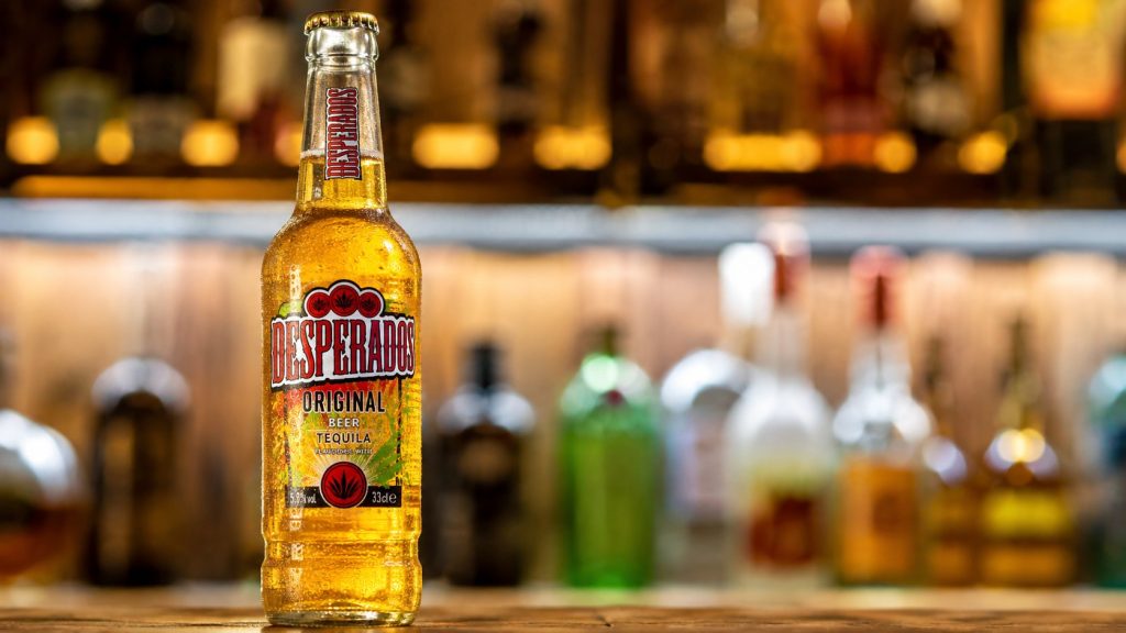 Desperados — piwo aromatyzowane tequilą — to weteran i wciąż jedna z najsilniejszych marek segmentu imprezowego. Charakterystyczna butelka z limonkową grafiką od lat broni swojej pozycji w lodówkach Żabki i dyskontów. Oto aktualne ceny Desperadosa na 2026 rok.